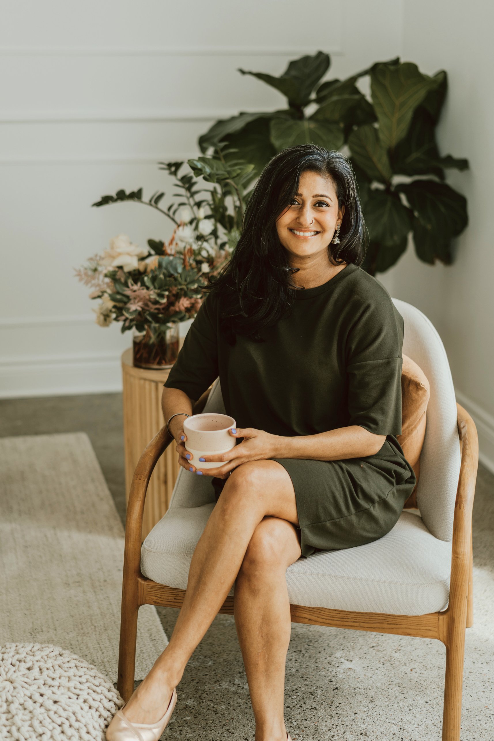 Sandeep is wearing a green dress sitting on a chair and smiling, holding a cup cup of coffee