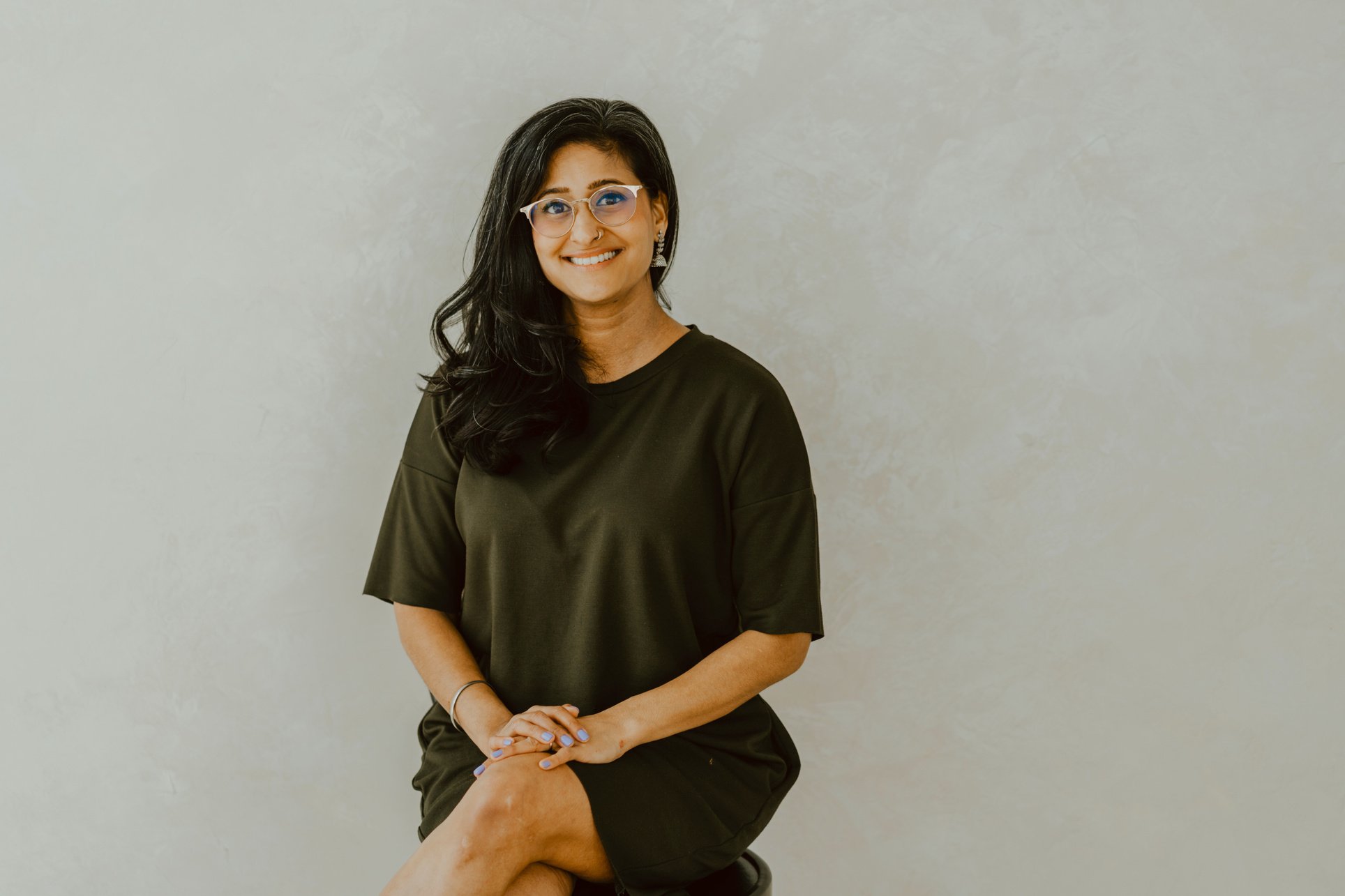 Sandeep is seated on a stool. She is smiling and wearing gold framed glasses and a green dress.