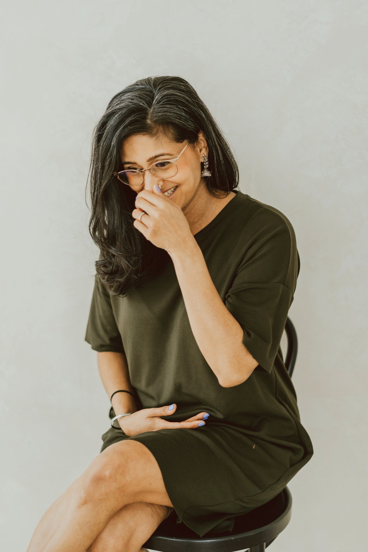 Sandeep is seated on a stool and wearing gold frame glasses and a green dress. She is laughing and covering her mouth with her hand.